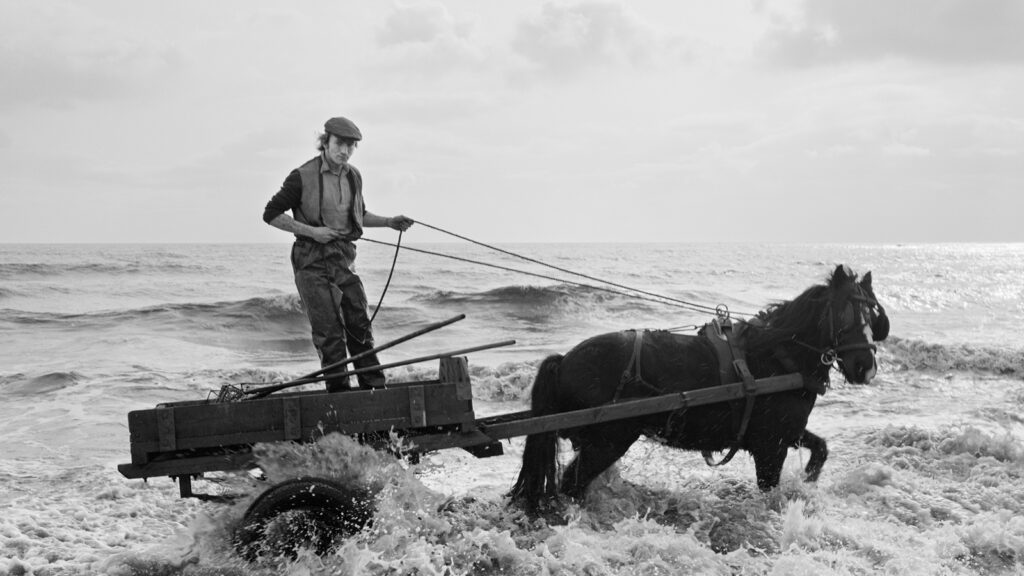 Das Foto zeigt einen Mann, der auf einem Wagen steht und ein Pferd durch das Wasser führt. Das Bild stammt von Chris Killip