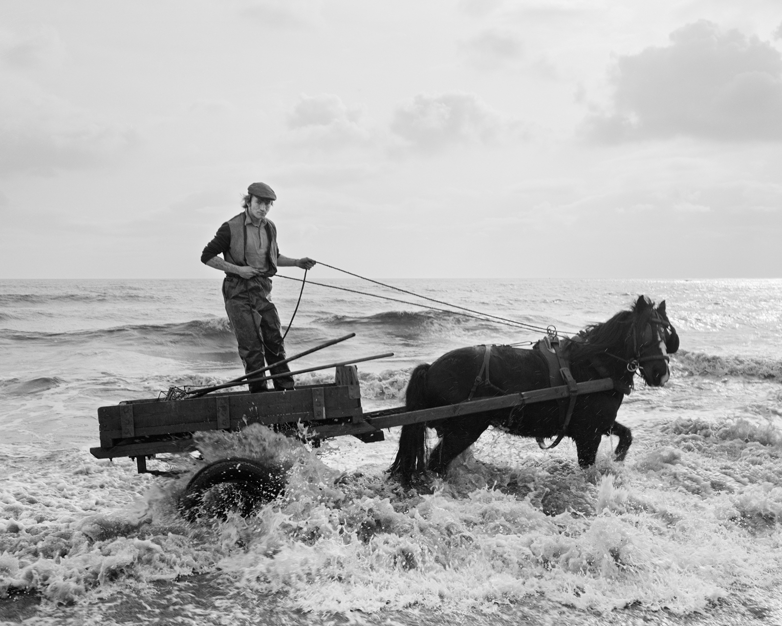 Das Foto zeigt einen Mann, der auf einem Wagen steht und ein Pferd durch das Wasser führt. Das Bild stammt von Chris Killip
